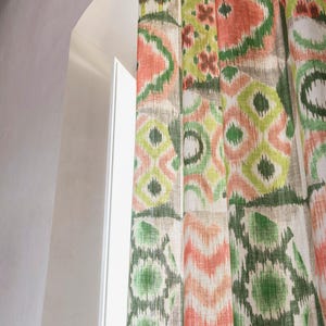 Könnte beinhalten: Vorhänge mit farbenfrohem geometrischem Muster in Korallen-, Grün- und Grautönen. Der Stoff ist in vertikalen Falten drapiert, mit einem Fenster und einer weißen Wand im Hintergrund. Die Vorhänge verleihen dem Raum eine lebendige Note.