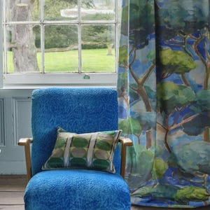 May include: A blue upholstered armchair with wooden arms and a patterned pillow. The pillow has a green and gray geometric design. Behind the chair is a window and a curtain with a tree-themed painting.