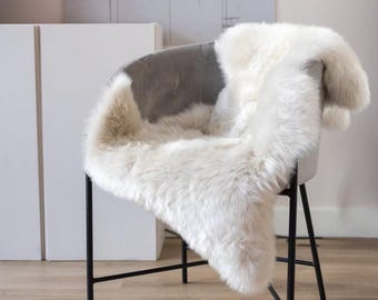 May include: A plush, off-white sheepskin rug draped over a gray, curved-back chair with thin, black metal legs. The long-haired, textured fleece covers the seat and backrest, creating a soft, layered appearance against the chair frame.