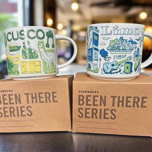 Puede incluir: Dos tazas Starbucks "Been There Series", una con la etiqueta "Cusco" y la otra "Lima", se asientan sobre cajas marrones. La taza de Cusco presenta ilustraciones verdes y amarillas. La taza de Lima tiene diseños azules y verdes.