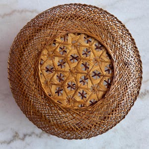 May include: A round, woven basket with a natural brown color. The basket features a detailed, geometric pattern in the center and a woven border. The basket is made of thin strips of wood, creating an open, airy design.
