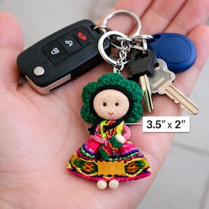 May include: A doll keychain measuring 3.5 inches by 2 inches, featuring a fabric face with black eyes and a stitched mouth, green crocheted hair, and a colorful patterned dress with a yellow accent. It is attached to a metal ring with keys.
