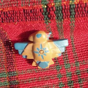 May include: A small, bird-shaped figurine with a tan body and blue wings and accents. The bird has a sunburst design on its chest. The figurine is set against a red and green plaid background.