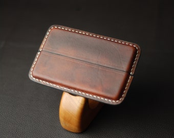 May include: A brown leather card holder with white stitching around the edges. The holder has a single seam down the center.