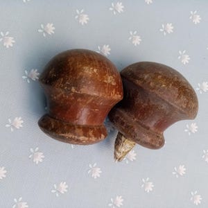 Puede incluir: Dos tiradores de armario antiguos de madera con acabado marrón oscuro. Los tiradores tienen una parte superior redondeada y una base acampanada, que muestra signos de desgaste. Se colocan sobre un fondo azul claro con un estampado floral.