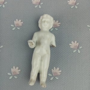 Peut inclure: Petite figurine en porcelaine blanche représentant un enfant, avec un bras manquant. La figurine a un visage détaillé et est debout. Le fond est un tissu bleu clair avec un motif floral.