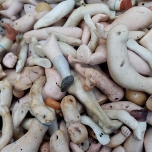 May include: A close-up of a pile of antique doll limbs, including arms, legs, and torsos. The porcelain pieces are in shades of pink, white, and tan, with signs of age. The limbs are scattered, creating a textured, close-up view of the doll parts.