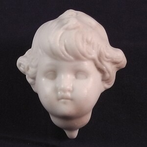 May include: A white porcelain sculpture of a child's head with curly hair. The sculpture is detailed with realistic features, including eyes, nose, and mouth.