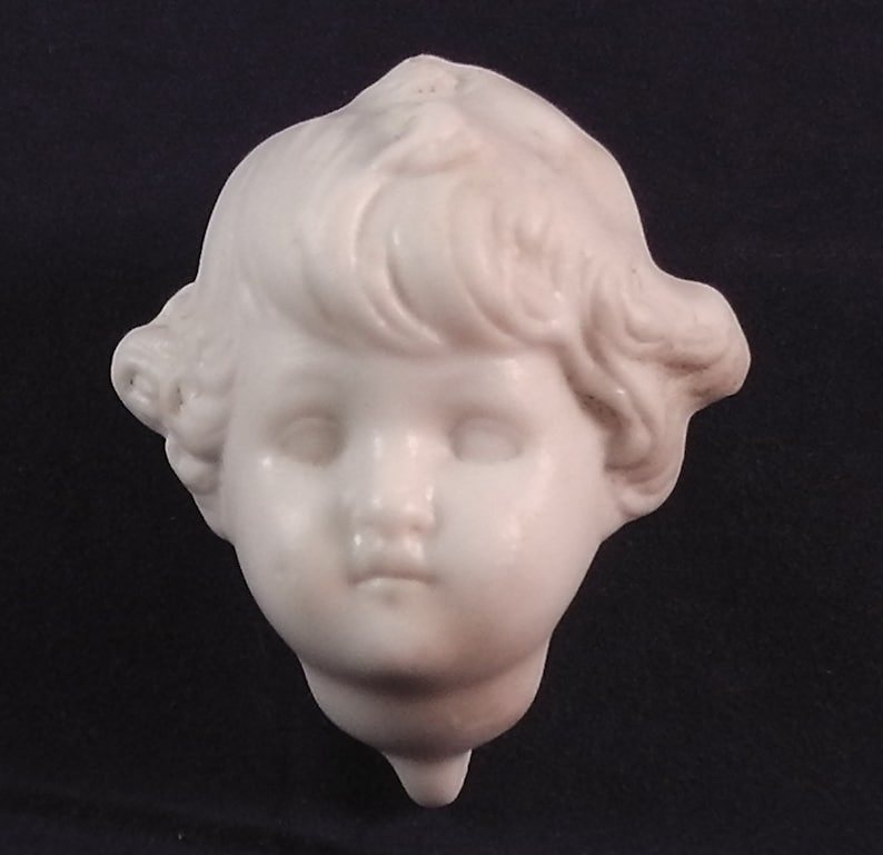 May include: A white porcelain sculpture of a child's head with curly hair. The sculpture is detailed with realistic features, including eyes, nose, and mouth.