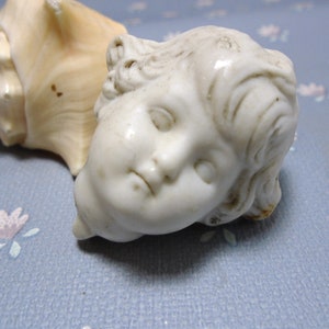 May include: A white porcelain cherub figurine with closed eyes and a delicate, sculpted face. The cherub is lying on its side, with its head resting on a seashell.