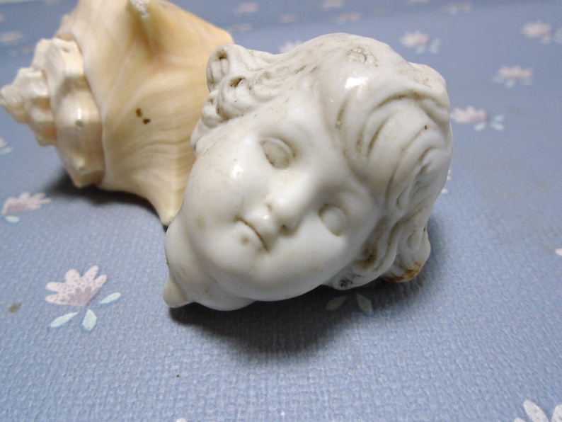 May include: A white porcelain cherub figurine with closed eyes and a delicate, sculpted face. The cherub is lying on its side, with its head resting on a seashell.