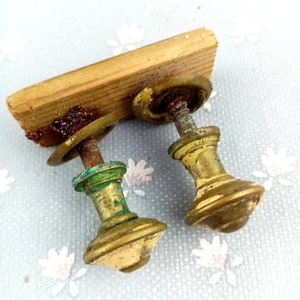 May include: Two antique brass drawer pulls with a wooden backing. The pulls have a tarnished gold finish with green oxidation and a decorative, rounded design. The wooden backing is a light brown color and shows signs of age.