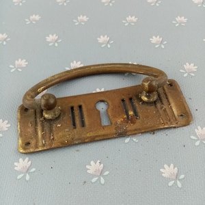 Könnte beinhalten: Ein antiker Schubladengriff aus Messing mit gebogenem Griff und rechteckiger Rückplatte. Die Rückplatte hat ein Schlüsselloch und vertikale Schlitze. Die Hardware ist gealtert und weist eine Patina auf.
