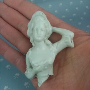 May include: A small, off-white porcelain figurine of a woman with flowing hair and a draped bodice. The figurine has one arm raised and is likely a decorative item or part of a larger set. The details are delicate, and the piece has an aged appearance.