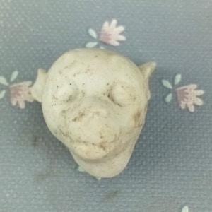 Könnte beinhalten: Ein weißer Porzellandollenkopf mit geschlossenen Augen und leicht geöffnetem Mund. Der Dollenkopf hat eine strukturierte Oberfläche und scheint vintage zu sein.