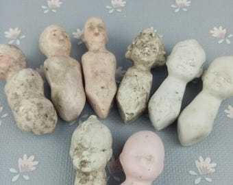 May include: A collection of nine vintage porcelain doll heads in various shades of white, pink, and gray. The doll heads are all different sizes and shapes, and some have painted facial features.