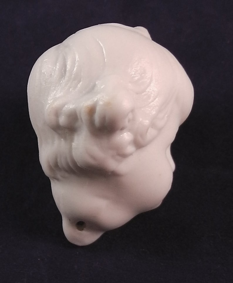 May include: White ceramic figurine of a person's head with long hair. The figurine is smooth and has a hole in the bottom.