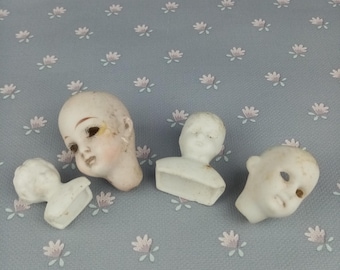 May include: Four porcelain doll heads, each with painted facial features. The heads are detached and some are on small bases. The heads are white with painted details.