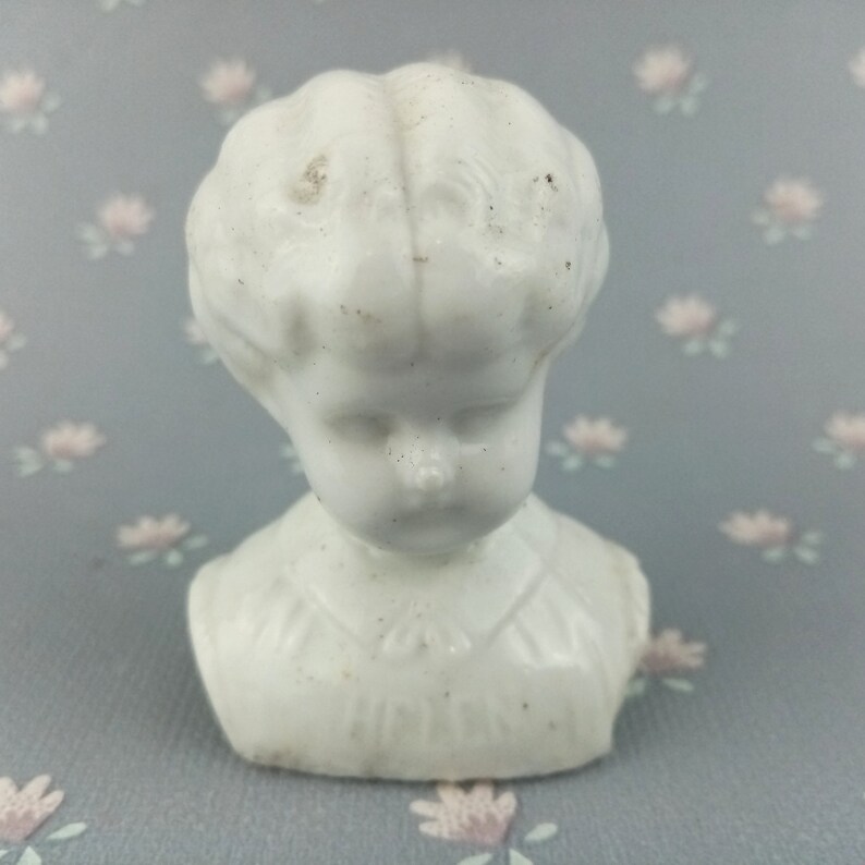 Op de afbeelding: Witte porseleinen poppenkopbuste met een eenvoudige witte jurk. De pop heeft een lieve uitdrukking met gesloten ogen en een kleine mond.