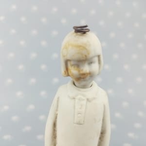 以下が含まれることがあります： ボブヘアの人物をかたどったオフホワイトの小さな陶器製フィギュア。頭頂部には茶色のワイヤーの輪が付いています。襟付きのシャツを着ており、前立てにはボタンのディテールがあります。顔には茶色の変色が見られます。