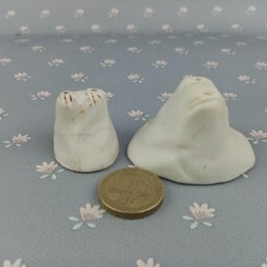 May include: Two white ceramic salt and pepper shakers shaped like heads. The shakers are on a light grey background with a floral pattern.