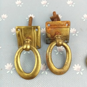 May include: Two vintage brass drawer pulls with oval handles and rectangular backplates. The pulls have a tarnished, aged appearance, with visible wear and tear. The screws are attached to the backplates. The background is a light blue fabric with a floral pattern.