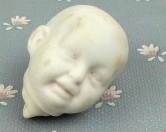 May include: A white porcelain doll head with closed eyes and a slightly open mouth. The head is slightly dirty and has a textured surface.