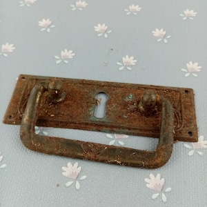 May include: A vintage, rectangular drawer pull with a keyhole and a curved handle. The metal hardware has a distressed, rusty brown finish with hints of verdigris. Decorative floral patterns are etched into the metal.