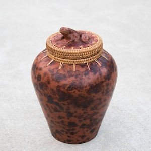 May include: A brown ceramic jar with a woven lid featuring a frog sculpture. The jar has a speckled brown and black pattern.