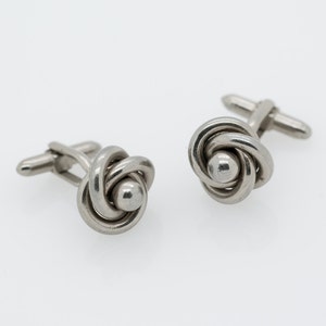 May include: A pair of silver cufflinks with a knot design. The knots are made of intertwined metal and have a small, round bead in the center.