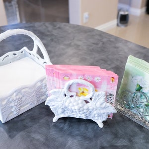 Puede incluir: Tres servilleteros decorativos. Un servilletero blanco de cerámica con forma de cesta con asa, un servilletero blanco con diseño de uvas y un servilletero de cristal transparente. Las servilletas son rosas y verdes con diseños florales.