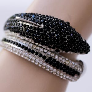 May include: A set of two bracelets, one black and one silver, adorned with sparkling crystals. The black bracelet is covered in small, black crystals, while the silver one features clear crystals. The bracelets are worn on a wrist.