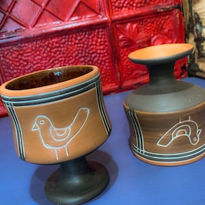Op de afbeelding: Twee bruine keramische bekers met een zwarte voet en een wit vogelontwerp. De bekers staan op een blauw oppervlak.