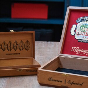 以下が含まれることがあります： 木製のシガーボックスが2つ。1つは赤い蓋に「Hemingway Reserva Especial」と書いてあり、もう1つは茶色の蓋に「Caesar Hand Made in Honduras」と書いてある。