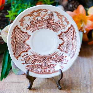 Könnte beinhalten: Ein weiß-brauner Zierteller mit einer detaillierten Illustration einer Hafenszene, einschließlich Schiffe und Gebäude. Der Teller hat einen gewellten Rand und wird auf einem Holzständer präsentiert. Der Text auf dem Teller lautet "New York Harbor Lands".