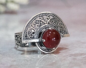 May include: A silver ring with a red carnelian stone set in a filigree design. The ring is shaped like a fan and has a delicate, intricate pattern.