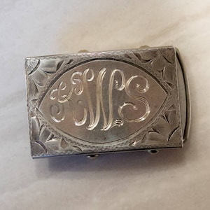 May include: A silver belt buckle with an engraved monogram of the letters "JLS" in a decorative oval shape.