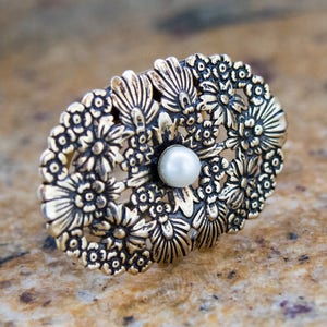 May include: An oval-shaped, gold-toned ring with a floral design. The ring features a central pearl and is surrounded by detailed, embossed flowers. The ring is set against a neutral background, highlighting its intricate details.