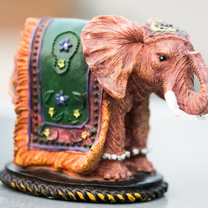May include: A small, decorative elephant figurine with a colorful patterned saddle. The elephant is made of painted resin and has a detailed, realistic design.