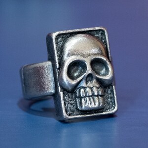 May include: A silver ring with a skull design. The skull is raised and detailed, with a rectangular frame.
