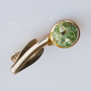 May include: A gold-toned tie clip featuring a circular, light green stone setting. The stone appears to be composed of smaller, irregular pieces. The clip has a simple, elegant design, suitable for formal wear.