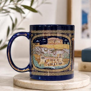 May include: A deep blue ceramic mug featuring a gold-toned Greek key border and a detailed illustration of the Parthenon in Athens. The design includes architectural ruins and the text "Parthenon of Athens" printed in a small, clear font.