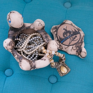 May include: A ceramic turtle trinket box with a black and white design. The box is open and filled with a pearl necklace and a silver necklace with a large pendant. The turtle box is sitting on a blue fabric surface.
