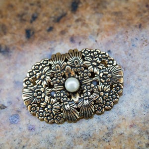 May include: Oval-shaped gold-toned brooch with a floral design. The brooch features a central pearl surrounded by detailed flowers and leaves. The metal has an antique finish, giving it a vintage look. A classic accessory.