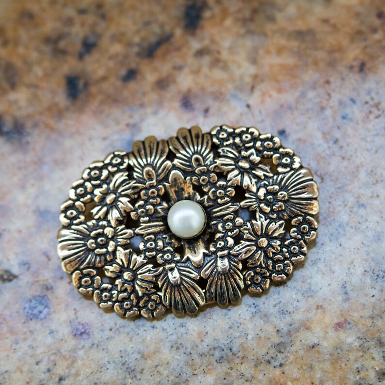 May include: Oval-shaped gold-toned brooch with a floral design. The brooch features a central pearl surrounded by detailed flowers and leaves. The metal has an antique finish, giving it a vintage look. A classic accessory.