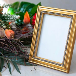 Könnte beinhalten: Ein goldener Bilderrahmen mit einem dekorativen Rand steht auf einem Tisch mit einem Blumenarrangement im Hintergrund.