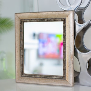 May include: A small, framed mirror with a gold-colored frame. The mirror is reflecting a blurred image of a colorful painting.