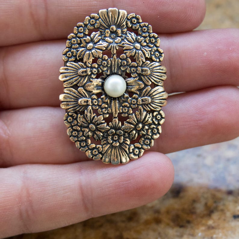 May include: An oval-shaped, antique gold-colored brooch with a floral design. The brooch features detailed flower and leaf patterns surrounding a central, round, white pearl. The brooch is held in a hand.