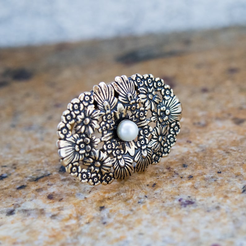 May include: An oval-shaped, gold-toned ring with a floral design. The ring features a central pearl and is surrounded by detailed, embossed flowers. The ring is set against a neutral background, highlighting its intricate details.