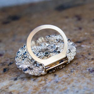 May include: A gold-toned scarf ring with an oval, floral-patterned base. The ring features intricate detailing and a small, green gemstone at the center. The ring is designed to hold a scarf or other fabric.